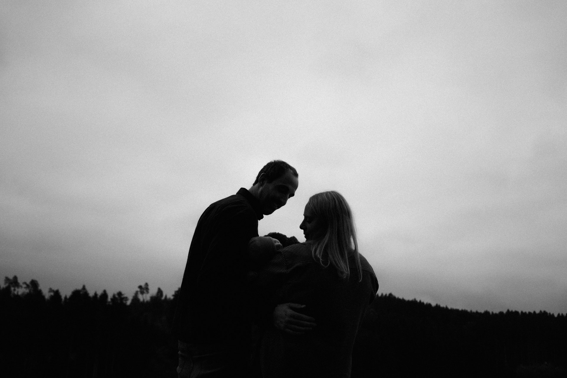Man erkennt bei diesem monochromen Foto nur die Silhouetten der jungen Familie; Vater und Mutter blicken auf ihren Sohn hinunter, im Hintergrund ist der Wald zu sehen. Das Foto ist im Rahmen der Newborn- und Familienfotografie entstanden.