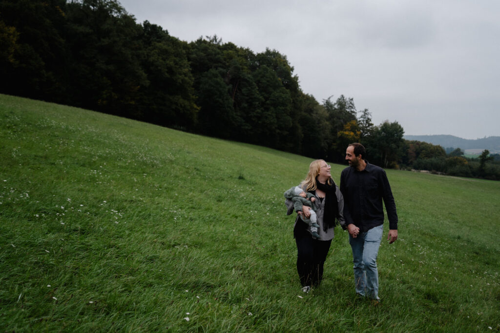 Vater und Mutter grinsen sich an, während sie mit ihrem Sohn in den Armen über eine Wiese spazieren. Das Foto ist im Rahmen der Newborn- und Familienfotografie entstanden.