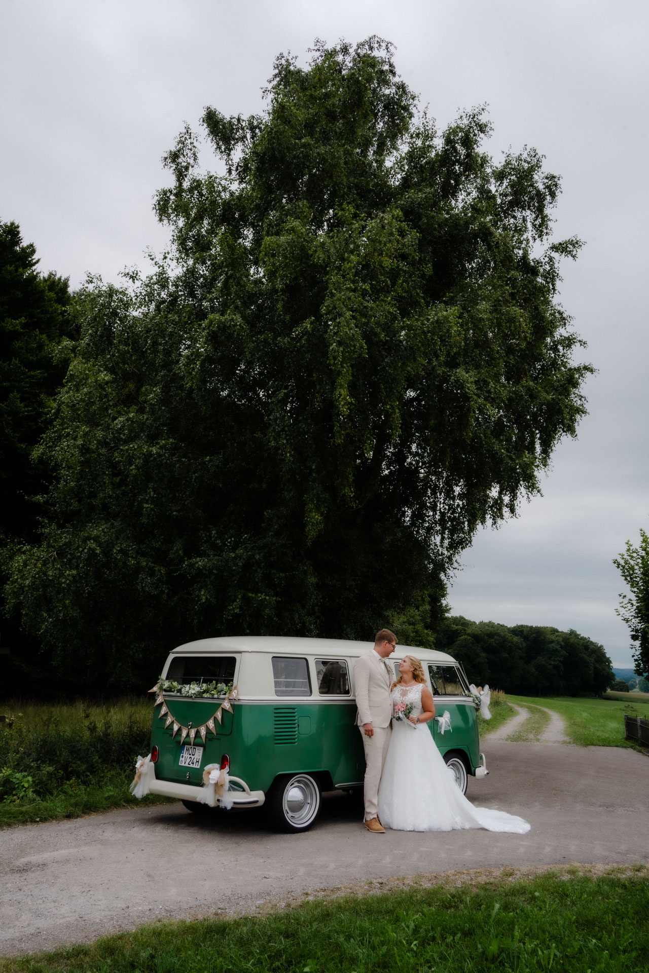 Das Hochzeitspaar posiert vor einem alten VW Oldtimer vor einem großen, schönen Baum.
