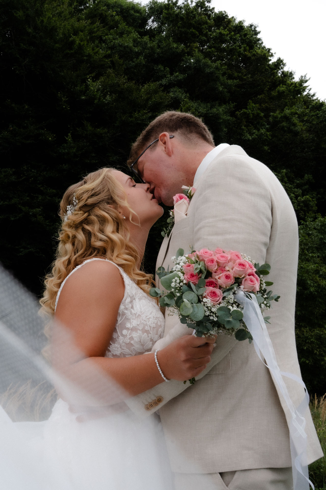 Braut und Bräutigam küssen sich vor dem Wald, ein Schleier verdeckt den Vordergrund ein wenig.