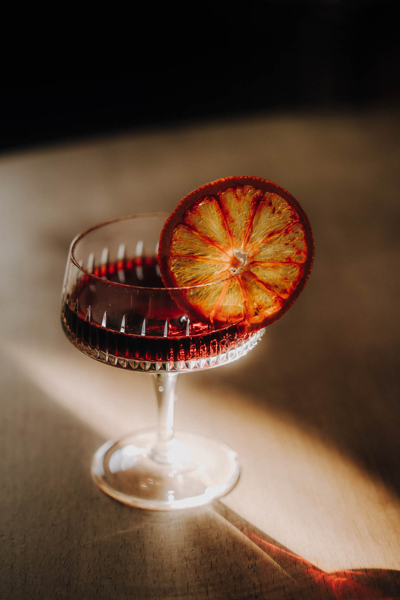 Das Foto eines roten Cocktails mit Orangenscheibe, erleuchtet von einem Sonnenstrahl und stilvoll arrangiert. Das Foto ist im Rahmen der Produkt-, Werbe und Foodphotography im Editorial Style entstanden.