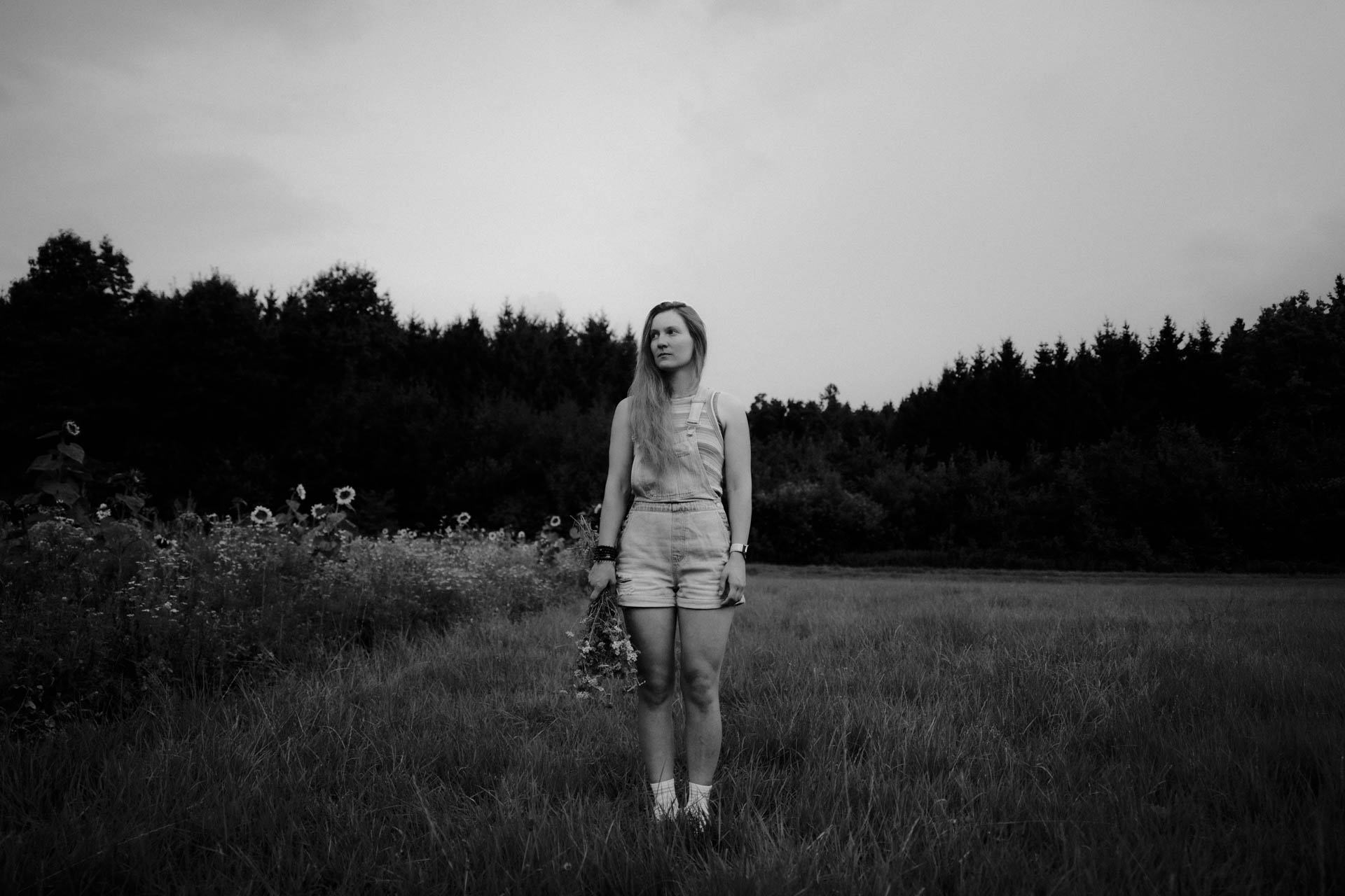 Das monochrome Foto einer Frau mit Blumenstrauß auf einer Wiese. Blumen und Baumreihen bilden führende Linien und machen das Foto stimmig, es ist im Rahmen der Portraitfotografie entstanden,