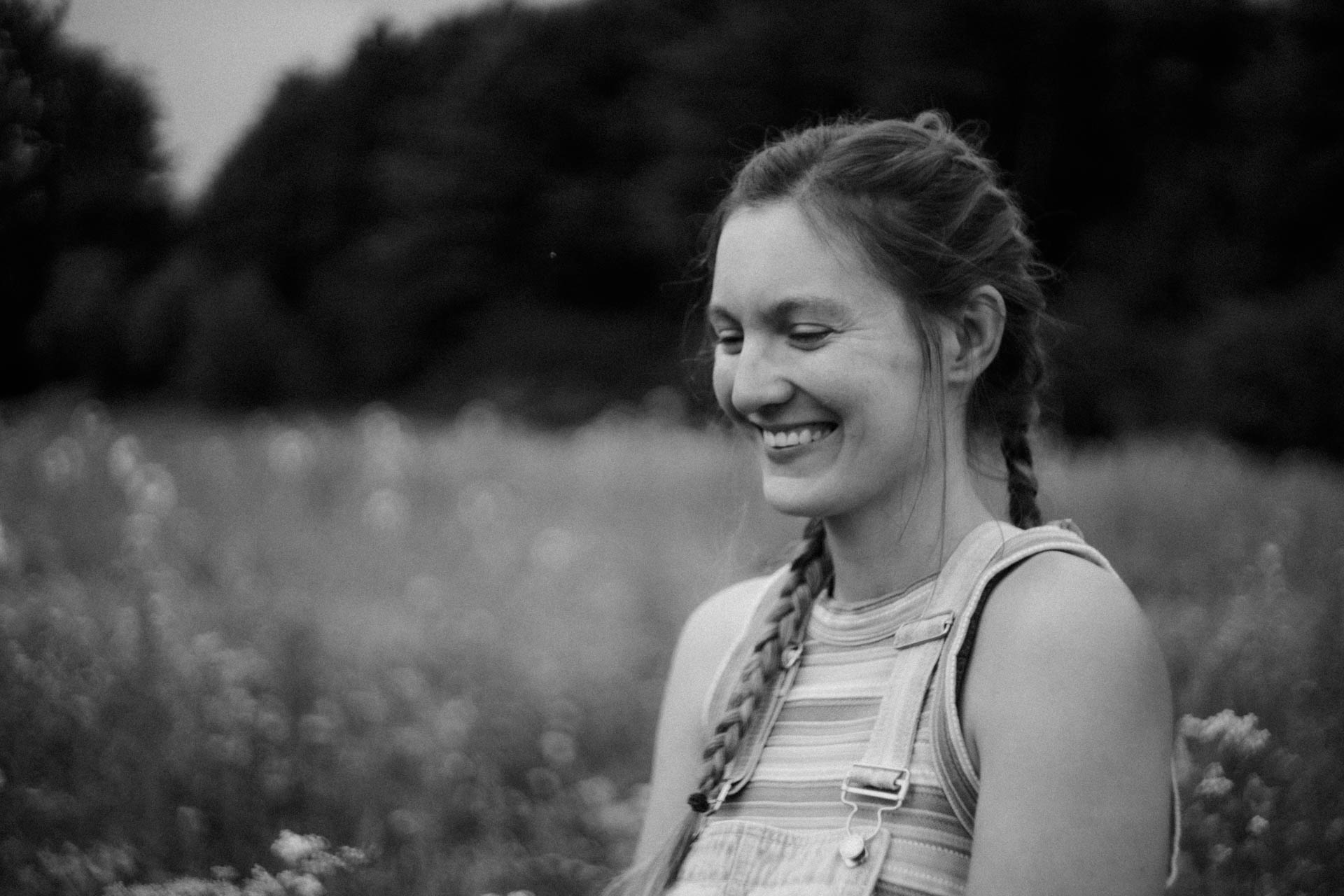 Das monochrom nostalgische Foto einer Frau mit Zöpfen, die in einer Blumenwiese sitzt, das Foto ist im Rahmen der Portraitfotografie entstanden.